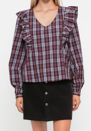 Kobieta ubrana w burgundową kratkowaną bluzkę z falbankami na ramionach oraz czarną spódnicę na guziki z sztruksu.