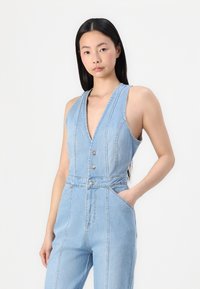 Combinaison en denim bleu clair avec un décolleté en V profond, design sans manches, boutonnière à l'avant, deux poches latérales et détails de couture verticaux.