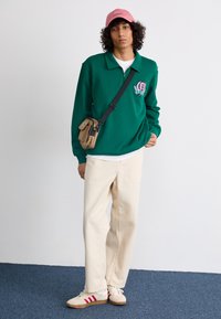 Grønn langermet polotrøye med glidelås på kragen, brodert logo, kremfargede vide bukser, og beige sneakers med røde striper. Rosa caps, crossover-veske.