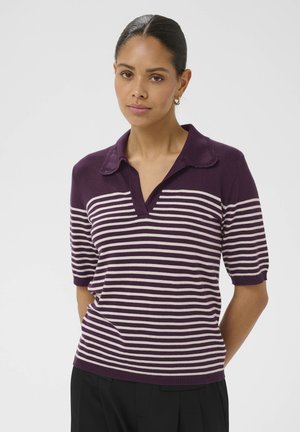 Kobieta o ciemnych włosach związanych w niski koczek, ubrana w fioletowo-białą koszulę w paski z krótkim rękawem i kołnierzykiem, stojąca z rękami za plecami.