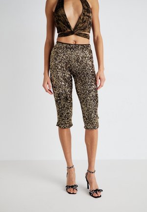 Kvinde iført sorte og gyldne paliet-cropped bukser, sorte høje hæle sandaler med perle-accenter og en brun fløjl halter top.
