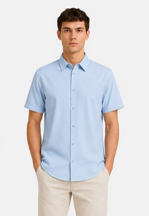 Junger Mann mit kurzen, lockigen dunklen Haaren, der ein hellblaues kurzärmeliges Hemd mit Knöpfen und beige Hosen trägt und die Hände in den Taschen hat.