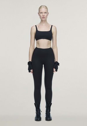 Une femme blonde se tient face à l’objectif devant un fond uni. Elle porte un soutien-gorge de sport noir, un legging, des gants sans doigts et des bottes à lacets.