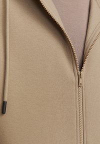 Beige zip-up hættetrøje med struktureret stof, der har en glat lynlås lukning og snore med sorte spidser.
