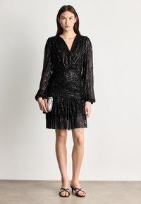 Vestido preto com lantejoulas, mangas longas, decote em V profundo, cintura franzida e barra com babados. Combinado com uma clutch prateada e saltos pretos.