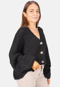 Frau in einer lockeren schwarzen Strickjacke mit großen goldenen Knöpfen und hellbeigen Hosen, vor einem schlichten Hintergrund stehend.