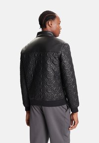 Chaqueta bomber negra con un patrón acolchado y acentos de cuero suave en los hombros, puños y dobladillo acanalados, combinada con pantalones de tonos neutros.
