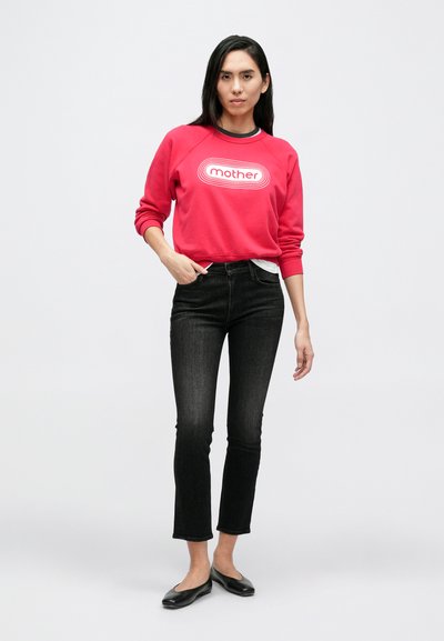 Raudonas džemperis su užrašu „mother“, juodos siaurėjančios džinsai ir juodi batai. Audinys atrodo minkštas; dizainas yra kasdienis su raukšlėtomis rankogaliais.