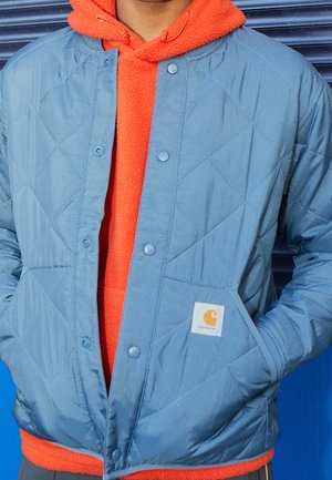 Person trägt einen leuchtend orangefarbenen Fleece-Hoodie unter einer hellblauen Steppjacke mit den Händen in den Taschen vor blauem Hintergrund.