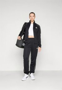 Svart atletisk joggedress med hvite striper, bestående av en jakke med glidelås og bukser med elastisk midje. Hvite joggesko og en svart duffelbag.