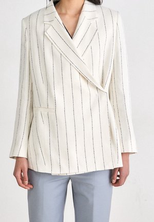 Femme portant un blazer blanc à fines rayures avec un devant croisé et un pantalon gris clair, debout devant un fond uni.