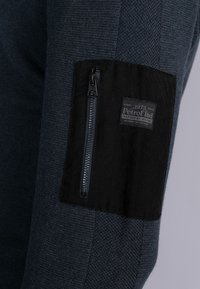 Marineblau gestrickter Stoff mit strukturiertem Muster, ausgestattet mit einem schwarzen Stoffeinsatz am Arm und einer Reißverschlusstasche, beschriftet mit "Petrol Ind."