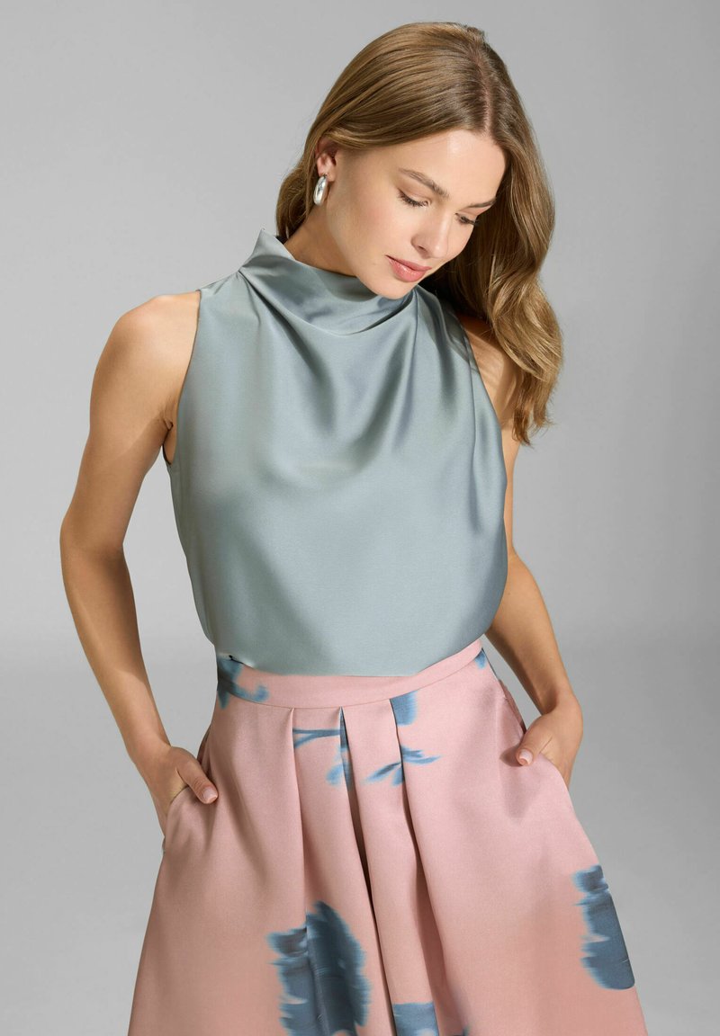Femme aux longs cheveux portant un haut sans manches en satin argenté et une jupe taille haute rose avec des motifs floraux bleus, les mains dans les poches, regardant vers le bas.