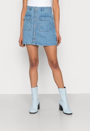 Femme portant une jupe en denim bleue à boutons sur le devant avec de grandes poches avant et des bottines bleu clair à talons, debout devant un fond uni.