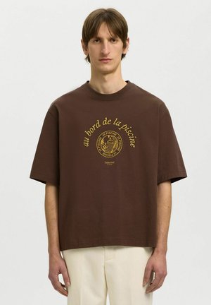 Joven con camiseta marrón extragrande con texto francés amarillo "au bord de la piscine" y gráfico, combinado con pantalones beige claro.