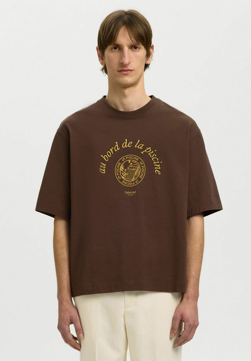 Jeune homme portant un t-shirt marron oversize avec le texte français jaune "au bord de la piscine" et un graphisme, associé à un pantalon beige clair.