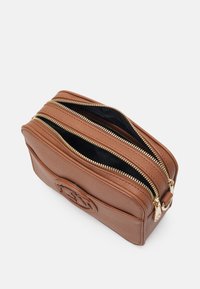 Brązowa skórzana torba z podwójnymi suwakami, teksturowana powierzchnia, czarne wykończenie wnętrza oraz wypukłe logo z boku. Kompaktowy i prostokątny kształt.