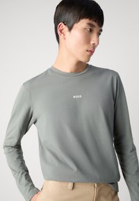 Langärmliges, rundhalsiges Shirt in gedämpftem Grün mit kleinem weißen "BOSS"-Logo auf der Brust und einer glatten, weichen Textur.