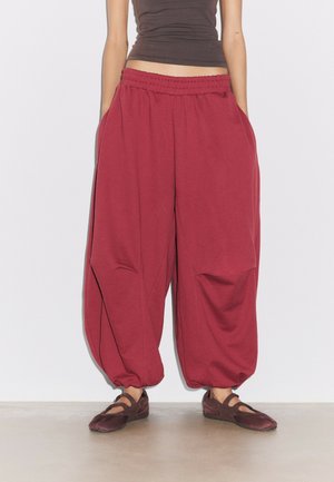 Personne portant un pantalon rouge ample avec des poignets élastiques et des chaussures plates marron, debout contre un mur blanc sur un sol lisse.