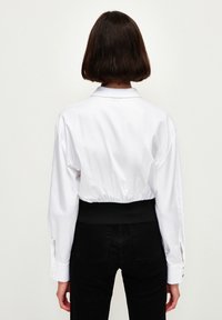 Maglietta bianca cropped con colletto, maniche lunghe e vita elastica. Presenta dettagli ai polsini e una texture liscia. Abbinata a jeans neri.