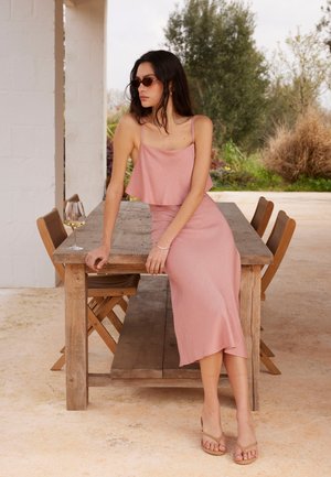 Kobieta w różowej sukience i okularach przeciwsłonecznych siedzi na drewnianym stole na zewnątrz z kieliszkiem wina i drewnianymi krzesłami wokół.