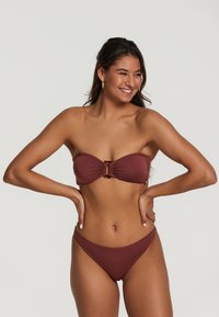 Bikini komplekts dziļā sarkanbrūnā krāsā ar bezsaru topu, kas izceļ priekšpuses rušie un dekoratīvo aparatūru. Atbilstoši zemas vidukļa bikini apakšdaļas.
