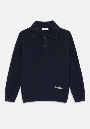 Sötétkék kötött pulóver bordás gallérral, két gombbal, hosszú ujjakkal, bordás mandzsettával és szegéllyel, valamint fehér hímzett "les deux" felirattal a szegély közelében.