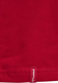 Gros plan sur un tissu rouge montrant un ourlet cousu avec une petite étiquette rectangulaire portant la mention « hummel » en texte blanc.