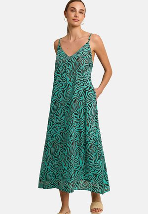 Femme portant une robe maxi turquoise sans manches avec un col en V, imprimé zèbre abstrait noir et des sandales beige tissées à enfiler.