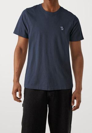 Navy cotton t-shirt with a crew neckline and short sleeves, featuring a small embroidered design on the chest. Paired with dark trousers.