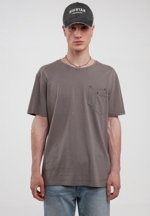 Grå bomulds t-shirt med en afslappet pasform, præget af en brystlomme og to trykknapper. Sammen med lyseblå denim jeans og en sort kasket.