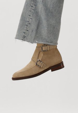 Bottes de cheville en suédine beige avec deux boucles décoratives, bout pointu et un petit talon marron. Portées avec un jean déchiré léger bleu.