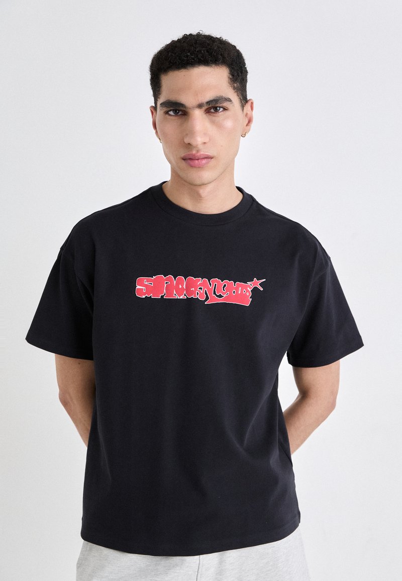 Tricou din bumbac negru cu un design grafic roșu, având text stilizat pe față. Mâneci scurte și un guler tip crew neck.