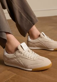 Beige Wildledersneaker mit weißen Akzenten, runder Zehenform, Schnürverschluss und strukturiertem Gummisohle. Sichtbare Nähte und Logo-Detail.