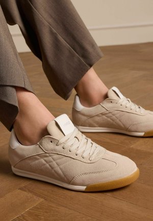 Beige suede sneakers with white accents, featuring a rounded toe, lace-up front, and textured rubber sole. Visible stitching and logo detail.