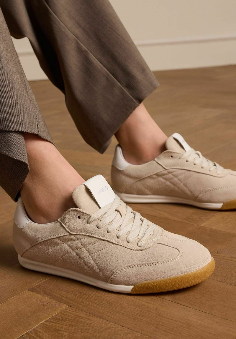 Beige Wildledersneaker mit weißen Akzenten, runder Zehenform, Schnürverschluss und strukturiertem Gummisohle. Sichtbare Nähte und Logo-Detail.