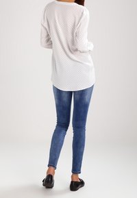 Blusa bianca a maniche lunghe con pois neri, abbinata a jeans skinny blu e scarpe nere flat, vista da dietro.