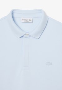 Vaaleansininen Lacoste-polo, jossa on kaulus, nappilista ja hienovarainen krokotiililogo kirjailtuna rintamukseen.