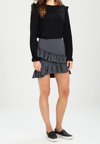 Schwarze Bluse mit langen Ärmeln und Rüschendetails, kombiniert mit einem schwarz-weiß gestreiften, gestuften Rock und schwarzen Slippern.