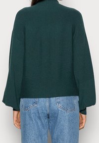 Personne portant un pull à manches longues vert foncé et un jean en denim bleu clair taille haute vue de dos sur un fond uni.