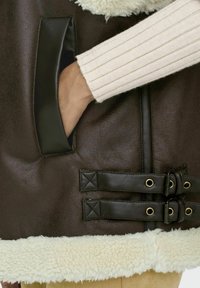 Bruine faux leren vest met een schapenwol trim, voorzien van een zak, twee gespsluitingen en een textuur detail.