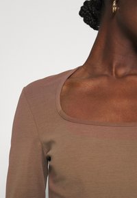 Långärmad tröja i en ljusbrun nyans, med rundad halsringning och mjukt, strukturerat tyg. Närbild av halsringningen och axelområdet.