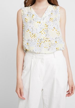 Mouwloze blouse met geel-grijze bloemenprint gecombineerd met witte hoge taille broek bij een staande persoon.