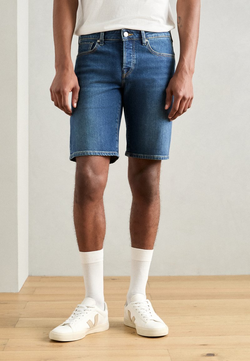Shorts en denim bleu foncé, avec une coupe classique et des ourlets retroussés, assortis à des chaussettes blanches et des baskets blanches.