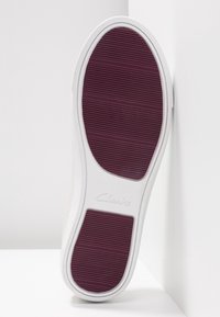 Valkoiset tennarit, joiden pohja on varustettu teksturoitulla burgundinpunaisella kumipinnalla, jossa on vaakasuorat urat ja kohokuvioitu "Clarks"-logo. Siisti muotoilu.