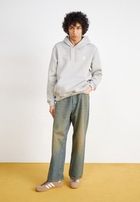 Felpa con cappuccio grigia con tasca frontale e logo bianco, abbinata a jeans larghi azzurri chiari e sneaker marroni con dettagli bianchi.