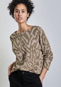 Beige Pullover mit einem abstrakten Muster in Beigetönen und Brauntönen, Rundhalsausschnitt, langen Ärmeln und gerippter Textur. Über dunklen Hosen getragen.