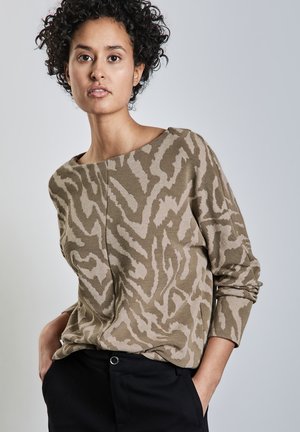Pull en beige avec un motif abstrait dans des nuances de beige et de marron, col rond, manches longues et texture côtelée. Porté par-dessus un pantalon sombre.