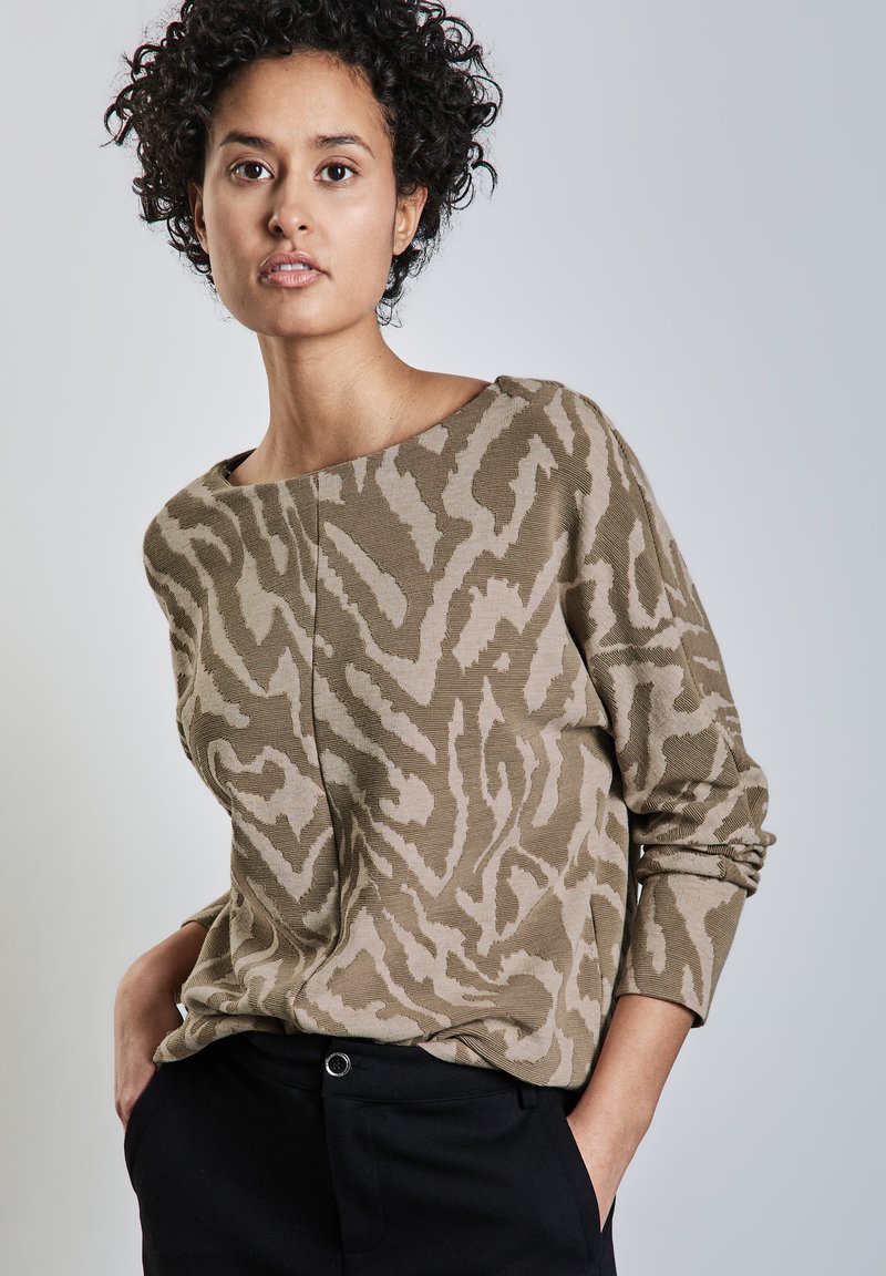 Beige Pullover mit einem abstrakten Muster in Beigetönen und Brauntönen, Rundhalsausschnitt, langen Ärmeln und gerippter Textur. Über dunklen Hosen getragen.