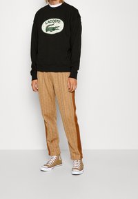 Sudadera negra con logo de Lacoste, pantalones marrones con patrón y franjas laterales, y zapatillas altas marrones con suelas blancas.